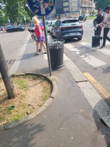 Curb cut onto a sidewalk that is blocked by a tree, a pole, and a trashcan. Milan, Italy near Milano Central Station